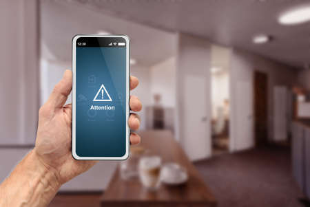 Male hand holding modern smartphone with security alarm app on screen over office backgroundの写真素材
