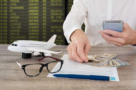 Man with smartphone applying travel insurance form in airport, plane model, glasses and cash money lying on tableの写真素材