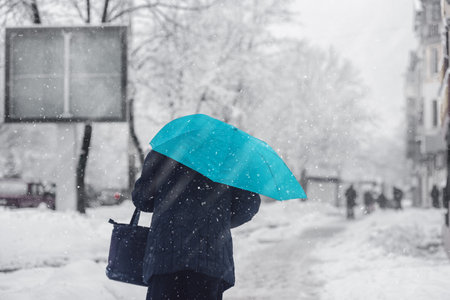 Back view of woman with blue umbrella walking on city street during heavy snowfall. Background: An elderly woman walking down a city lane in a heavy snowstorm clutching an azure umbrella.の写真素材