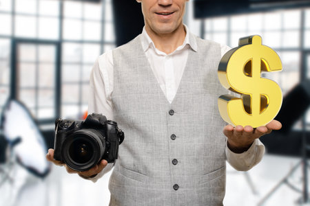Closeup of happy young man holding modern camera and dollar sign in photo studio, the essence of the...