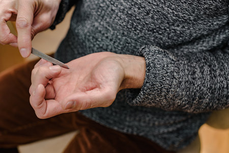 Closeup photo of adult man making manicure with steel nail file. Meticulous touch: A man focuses on his nails, displaying personal care and precisionの写真素材