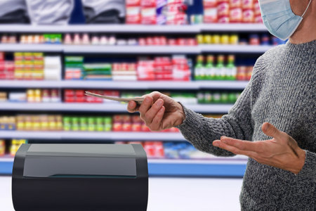 Closeup photo of man in medical mask paying at self-service counter using cash. Convenient shopping: Cash dollars utilized at self-service terminal for easy supermarket checkoutの写真素材
