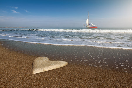 Stone heart lying on sandy beach with ship on ocean water background. Affection and fidelity symbolized by a heart-shaped stone on the sandy shore of the seaの写真素材