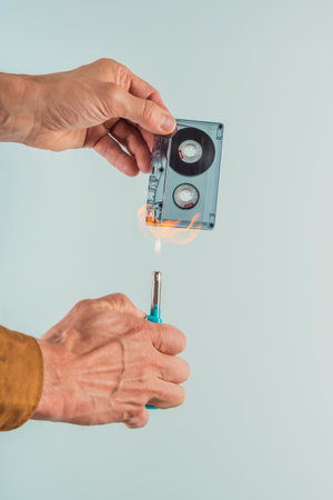 Male meloman burning old audio tape cassette on blue background. Audio cassette and lighter,...