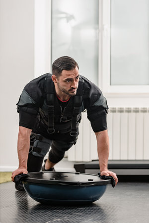 Young bearded man in electrical muscular stimulation suit exercising with bosu ball, Fitness goals: Electro stimulator in gym setting, ideal for promoting muscle developmentの写真素材