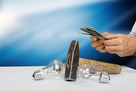 Closeup of man counting dollars money banknotes, hammer and light bulbs lying on table. Breaking incandescent bulbs with a hammer, this photo emphasizes the importance of replacing old lightingの写真素材
