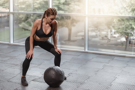 Strong fit slim woman working out with medicine ball, Crossfit zone gym: girl in black workout clothes, glass windowsの写真素材