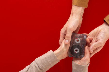 Flat lay photo of male and female hands holding retro audio cassette on red background. Close-up of hands with a retro audio cassette on a flat background, ideal for archive and nostalgiaの写真素材