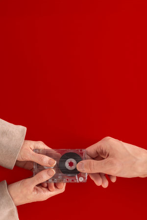 Closeup of man offering woman to listen to vintage audio cassette on red background. Close-up of hands passing an audio cassette on a flat background,の写真素材
