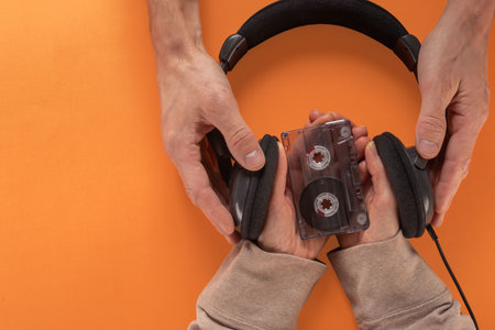 Female and male hands holding headphones and audio cassettes on orange background. Hands holding an audio cassette and headphones, flat lay, capturing the nostalgia of retro music playbackの写真素材