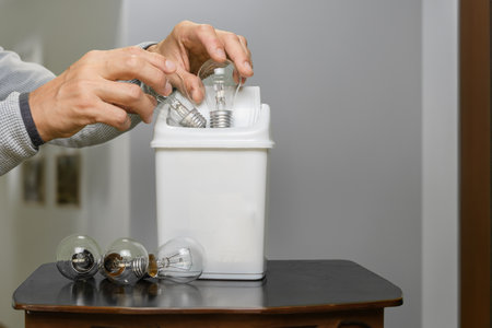 Male hand throwing light bulb into trash can at modern apartment. An incandescent lamp in a recycling bin within a stylish apartment, illustrating a commitment to sustainability and responsible livingの写真素材
