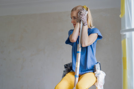 Unhappy young woman holding tool for repair and feeling fatigue after wall painting Woman feeling hopeless during a solo apartment renovation, emphasizing the struggles and failures of DIY home repairの写真素材