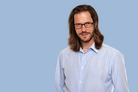 Portrait of young handsome man in glasses posing on blue background, Close-up portrait of a man with long hair and stylish glasses, posing against a bold color backgroundの写真素材