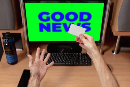 Excited young man with credit card looking at computer screen with message good news. Man at computer, keyboard visible, reacting positively to news on chromakey screenの写真素材