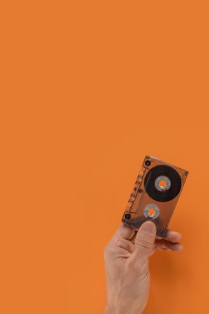 Unrecognizable man holding compact audio cassette on orange background, closeup photo. Hand gripping an audio cassette, showcased against a flat background for a nostalgic, retro feelの写真素材