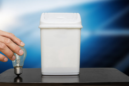 Man going to throw light bulb into trash bin as the symbol of recycle. An incandescent lamp being discarded into a trash can, set against a gradient blue background, symbolizing responsible recyclingの写真素材