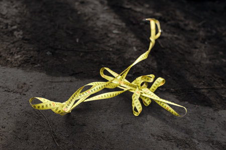 Yellow sewing tape measure lying on black ground, closeup photo. Old measuring tape twisted on ground, tool for DIY, landscaping or renovation projectsの写真素材