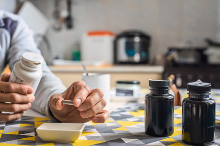 Adult man in home kitchen holding bottle of nutritional supplements, closeup photo. Man sitting at kitchen table holding supplement capsule, bottles and cup in backgroundの写真素材