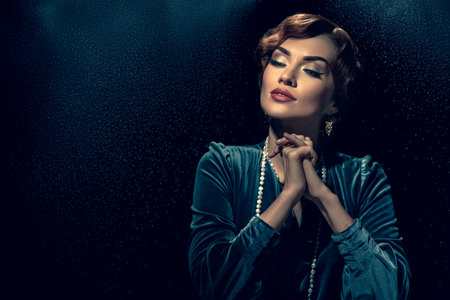 Romantic beautiful lady with retro wave hairstyle posing on dark background with dust particles. Pensive woman in retro dress with vintage hair and elegant jewelryの写真素材