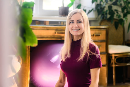 Portrait of beautiful smiling young woman practice yoga in light room. Relaxed woman in sportswear meditating indoors, bright room with natural light and plantsの写真素材