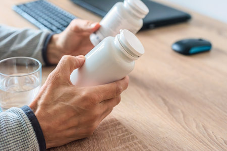Closeup of male hands examining jars with medication at home office. Workplace wellness, male hand holding vitamin jar beside computer keyboard and mouseの写真素材
