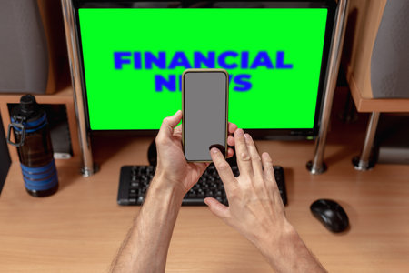 Male worker holding mobile phone with notification about money transfer. Man reading financial news on computer monitor, visible emotions, keyboard in viewの写真素材