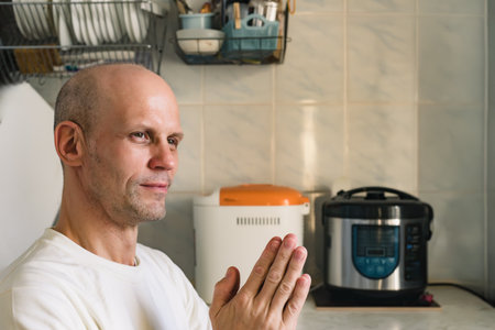 Young man baking bread at home and praying with hands together with hope expression on face. A man sits in the kitchen with his hands folded, waiting for food to cook, hoping to get a tasty meal.の写真素材
