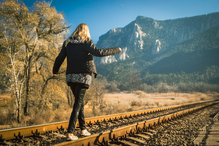 Happy blonde girl balancing walking on railway track with arms spread in mountain landscape. Girl balancing on train rails in scenic landscape with rocky cliffs and autumn trees outdoors.の写真素材