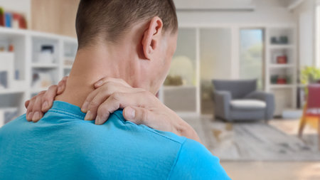 Young man with strong muscular ache massaging neck standing at home. Man holding neck in pain at...