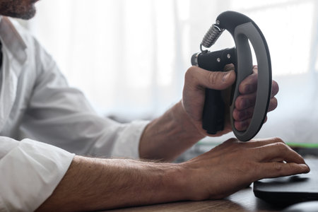 Male employee squeezing black hand gripper while working on computer. Close-up of hand training with...