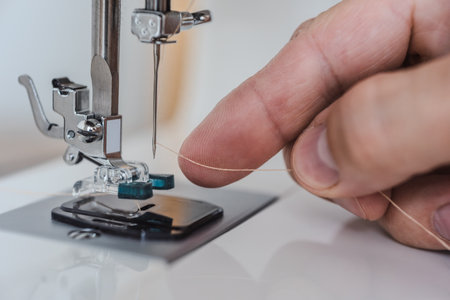 Male fingers putting thread in needle loop on electric modern sewing machine for start sewing. Manually threading sewing machine needle with fingers, close-up of precise processの写真素材
