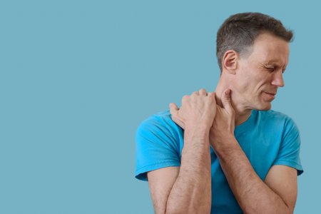 Portrait of young man feeling pain massaging stiff neck on blue background, man suffers from shoulder pain, touches muscle with grimace of discomfort.の写真素材