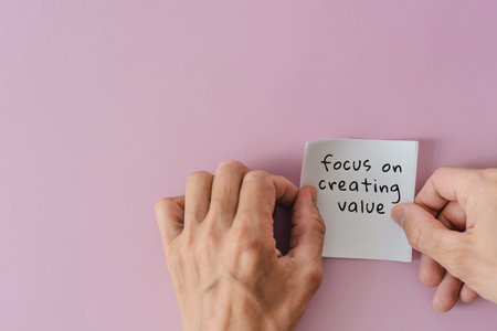 Focus on creating value text writing on sticky note against pink background. Hands placing note with...