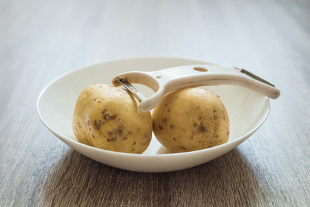 Raw potatoes and peeler in white bowl on wooden table. Potatoes and peeler in a bowl, simple preparation for home cookingの写真素材