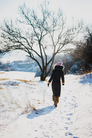 Pretty young woman walking among snowy trees on sunny winter day. Snowy walk in nature as a woman in warm winter clothes strolls alone, looking ahead in a peaceful momentの写真素材