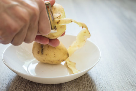 Chefs hand peeling a fresh and delicious potato for cooking, closeup photo. Home cooking process, peeling fresh potatoes into a white bowlの写真素材