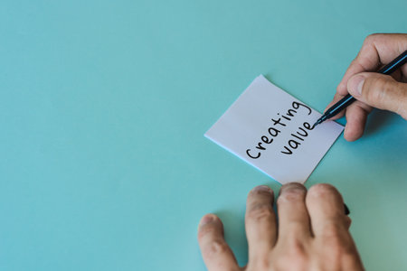 Male hands writing inscription "Creating value" on sticky note. Human hand writing strategic idea on pastel blue background, flat lay sceneの写真素材