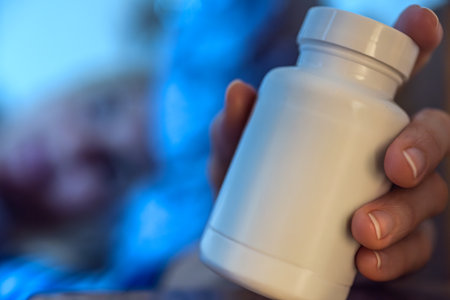 Closeup photo of woman holding bottle of dietary supplements or vitamins in hand. Hand holding white pill bottle with person resting in bed, concept of health, sleep or medicationの写真素材