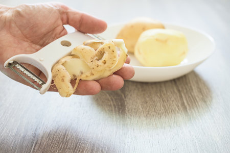 Man holding peeler and potatoes skin against bowl with peeled potatoes. Hand holding peeler and potato peel, close-up of kitchen preparationの写真素材
