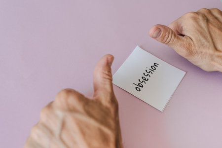 Man showing thumbs down and word "Obsession" written on sticky note. Paper note and two thumbs up on soft pink background, metaphor of praise and obsessionの写真素材