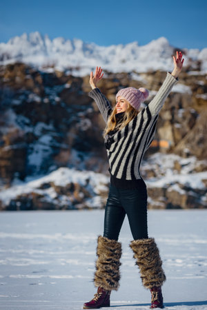 Young happy stylish girl with raised hands enjoying snow in mountains. Woman enjoying snow in the mountains, wearing a cozy sweater and boots. Full-length winter fun outdoorsの写真素材
