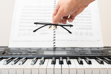 Male pianist holding glasses in front of music sheets, closeup photo. Hand holding glasses over keyboard and sheet music, concept of vision during piano learningの写真素材