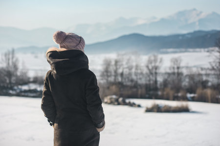 Woman tourist in mountains observing snow covered landscape. Woman in warm winter coat and knitted hat enjoying snowy mountain view from behindの写真素材