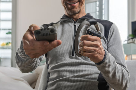 Young happy man holding expander in hand massaging muscles and watching TV. Smiling man using grip strengthener while watching television on couchの写真素材