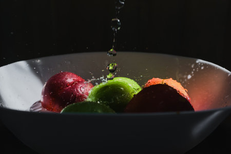 Washing fresh apples in streaming water on black background. Droplet of water falling on mixed fruits in bowl, splash captured in motionの写真素材
