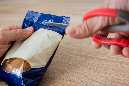 Man unpacking instant cocoa with scissors, closeup photo. Cutting food package with red kitchen scissors, close-up hands and bagの写真素材
