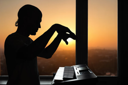 Silhouette of man sitting next to window at home and playing synthesizer at sunset background. Expressive piano hands silhouetted at sunset, home music practiceの写真素材