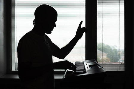 Silhouette of professional male musician playing synthesizer at home. Musician silhouetted by window...
