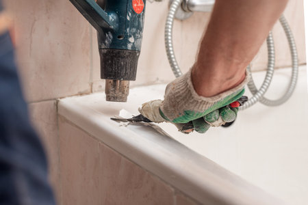 Closeup photo of man using heat gun and special knife to remove old sealant from bath. Peeling old bathtub coating with heat gun and scraperの写真素材