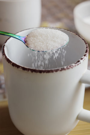 Close view shows iridescent teaspoon pouring white granulated sugar into white ceramic mug, with crystals spilling over rim and falling into cup under soft natural light, shallow depth, neutral